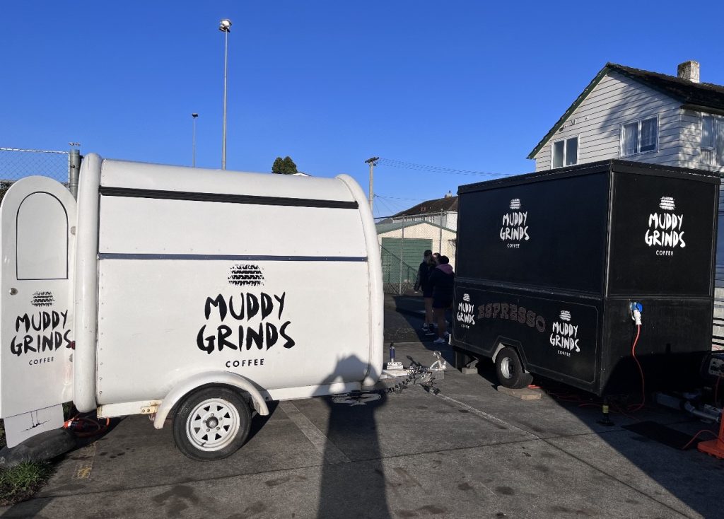 Photo of a small white trailer that has the logo Muddy Grinds on it and a black trailer with the same logo on it